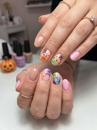 Cute monster Halloween nails with colorful cartoon faces, pastel pink polish, small spider web details, and playful spooky nail art designs